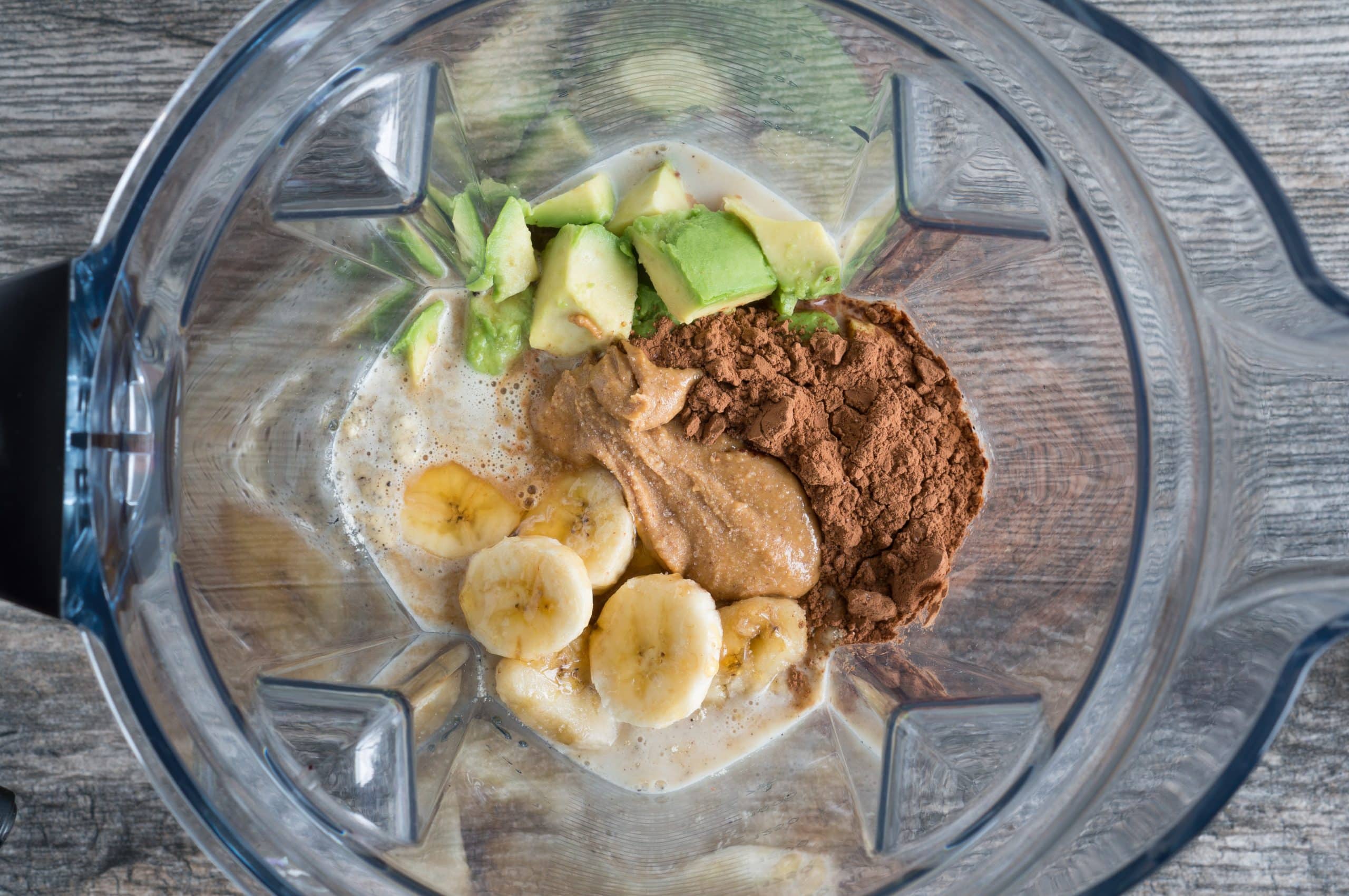 top down view of a clear blender cup filled with sliced bananas, peanut butter, cocoa powder and avocado